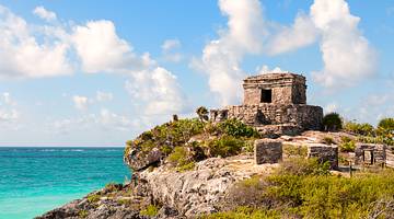 Ancient Mayan ruins on a rocky hill with green shrubs and the blue ocean behind