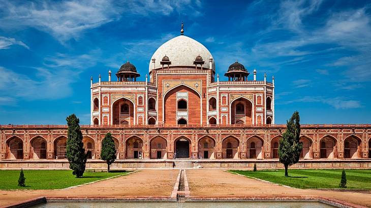 A pathway leading to a sandstone mosque with a white dome under a partly cloudy sky