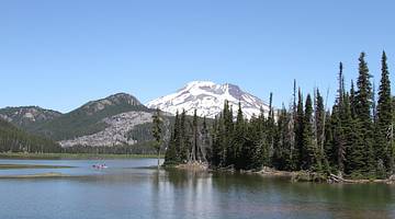 A snow-capped mountain at the back with water and pine trees in front