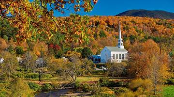 A white country church surrounded by fall foliage with a greenery-covered hill behind