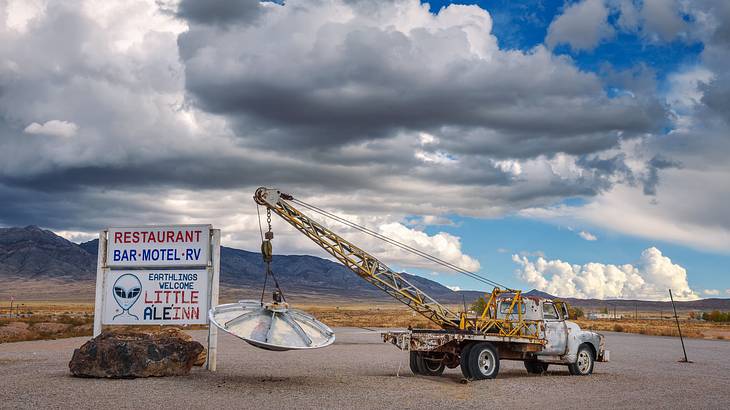A vintage pickup truck with a UFO beside a white signboard under a partly cloudy sky