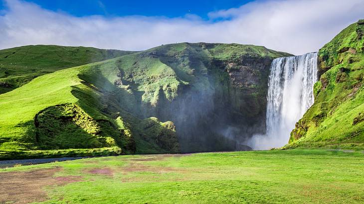 A waterfall cascading down a grass-covered hill under a blue sky with white clouds