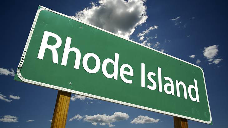 A green and white "Rhode Island" road sign with a blue sky and white clouds behind