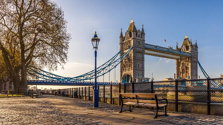 A bench on a cobblestone river walk with a view of a bridge under a clear sky