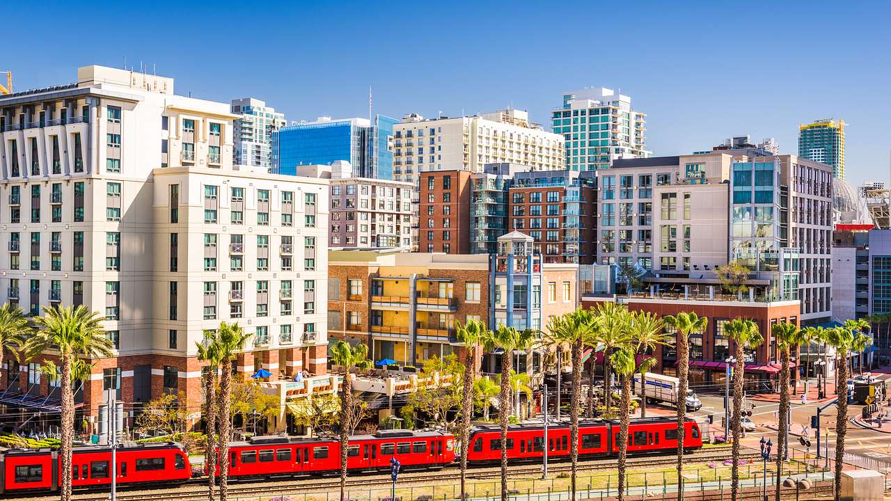 A red train going through a city with many buildings and palm trees