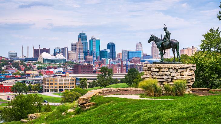 A statue of a horse and rider on a stone base against a downtown skyline