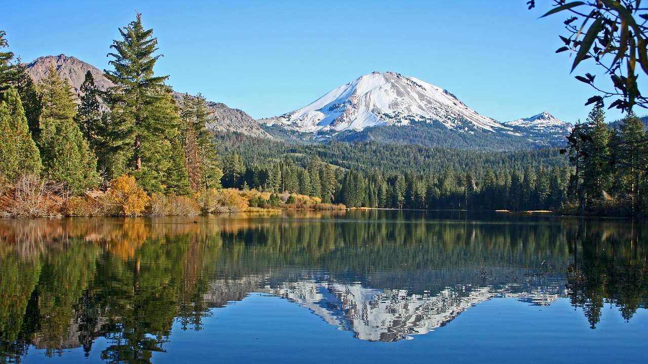 Snow-covered mountain and trees at its foot reflected over a lake on a clear day