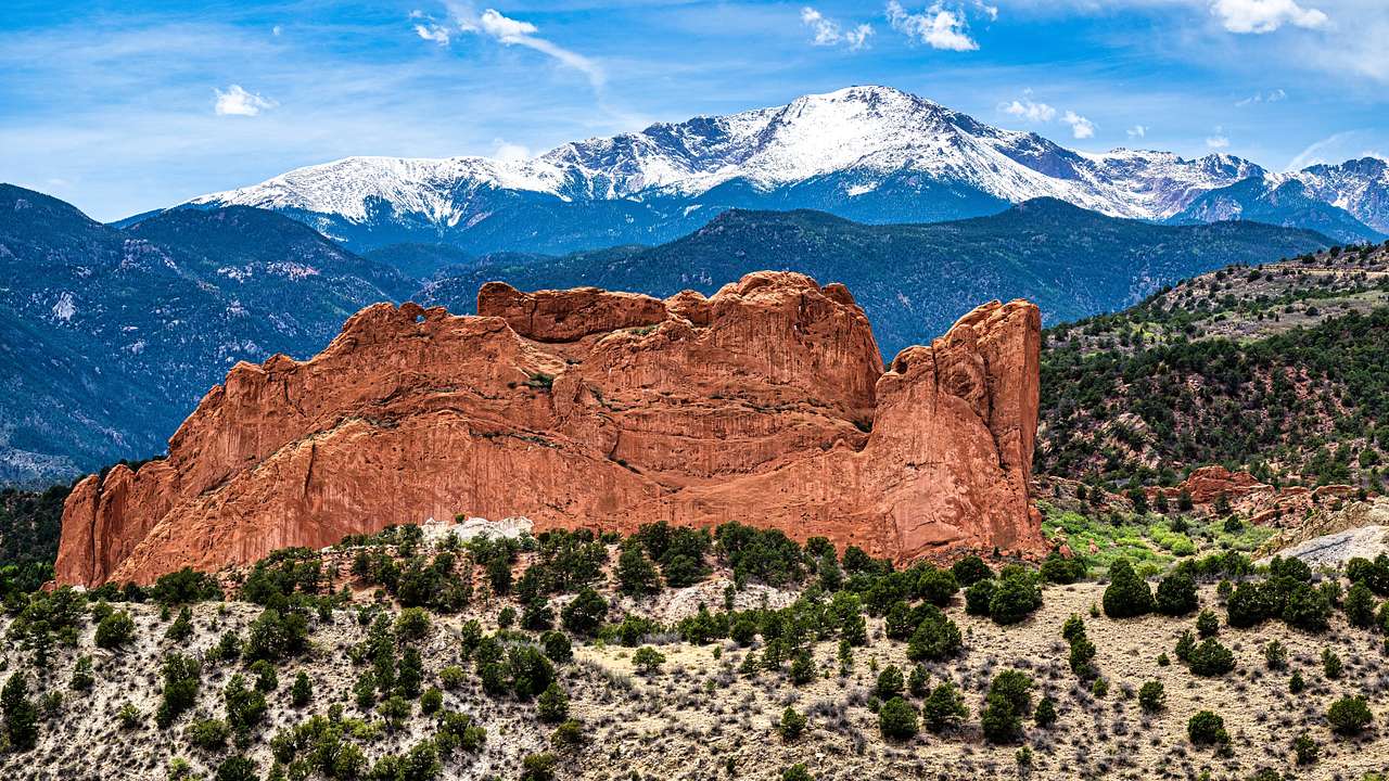 A wide red rock formation beside shrubs with a snow-capped mountain in the background
