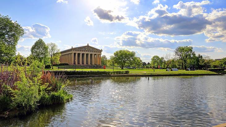 Parthenon building near a body of water, green grass, trees, and blue sky.