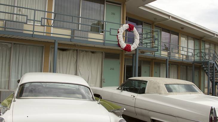 Two-story, older motel with balcony and two cars parked in front, cloudy sky