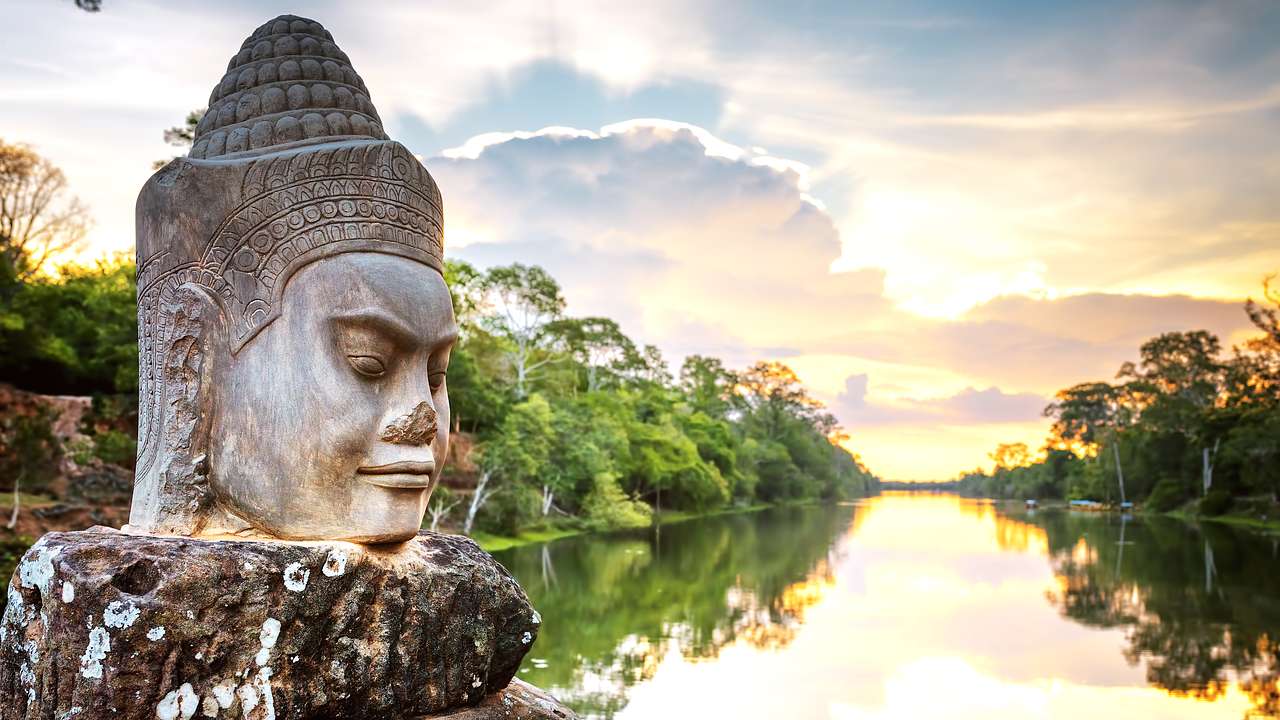 A stone-faced statue on a pathway overlooking water and trees at sunset
