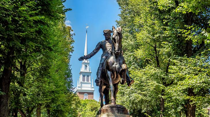 A statue of a man on a horse with a white church steeple and trees in the background