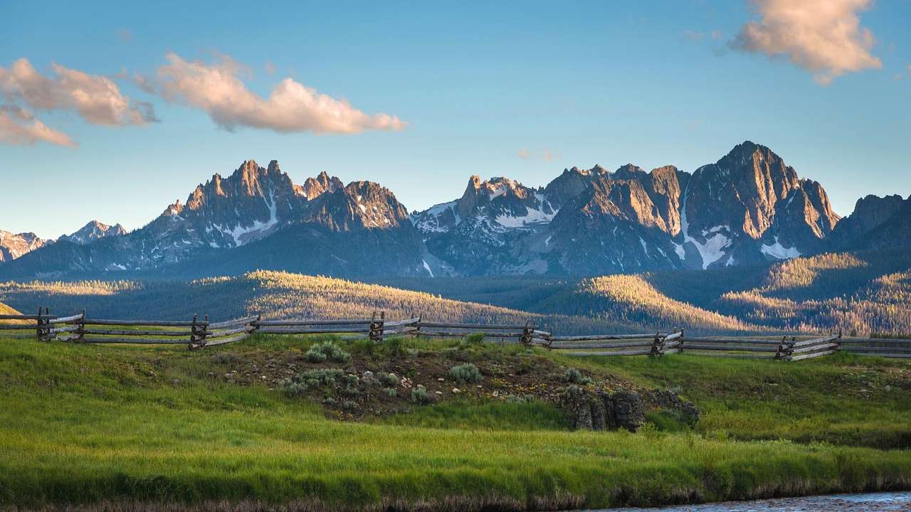 A gray mountain range with patches of snow, overlooking green grass lined by a fence