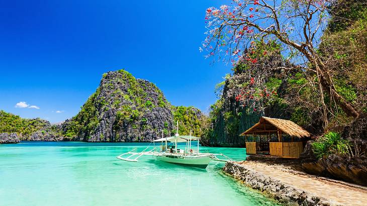 Turquoise water with a white boat on it next to a small hut and cliffs with greenery