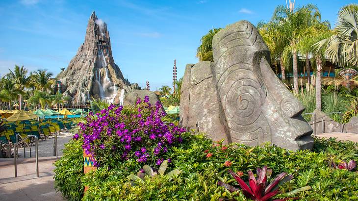 A rock statue of a human head surrounded by plants and a water-gushing peak behind it