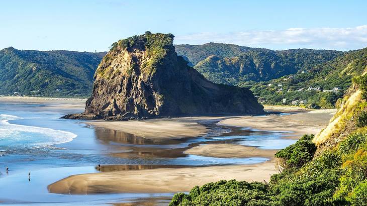 A large rock with greenery surrounded by water, sand, and green hills