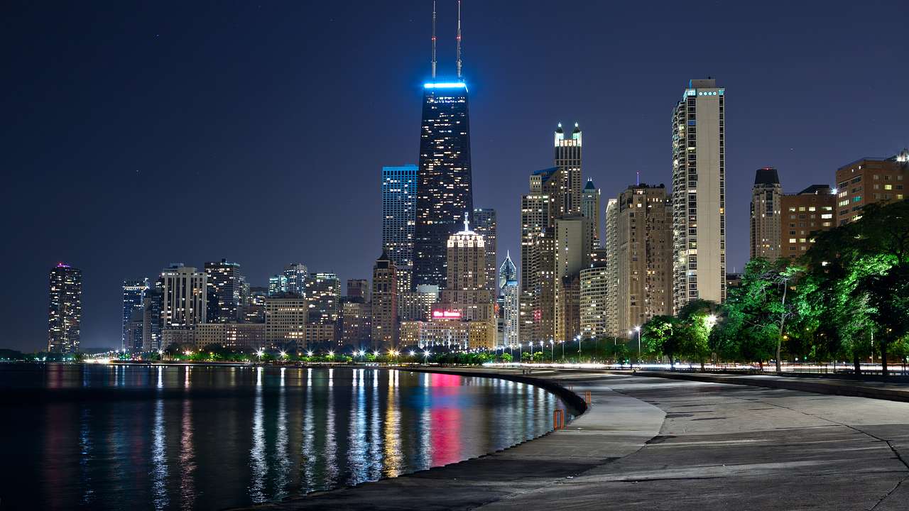 A lit-up skyline of modern buildings overlooking a pathway by a river