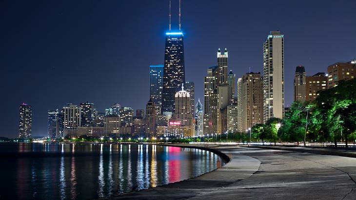 A lit-up skyline of modern buildings overlooking a pathway by a river A lit-up skyline of modern buildings overlooking a pathway by a river