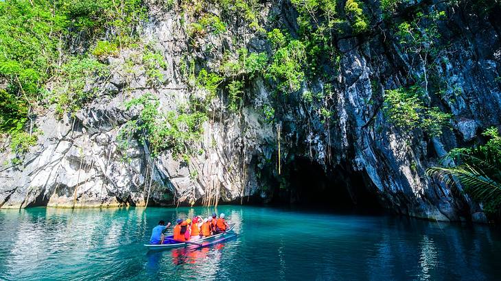 A river with a boat and people on it, next to a rocky cliff with a cave carved in it