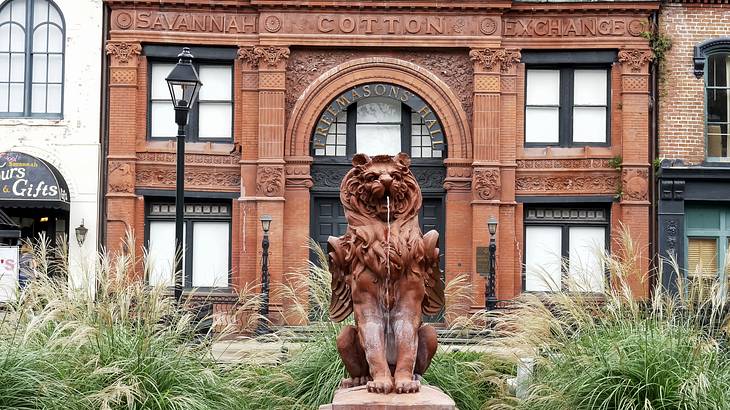 A brown lion statue surrounded by greenery, and a historic brown building behind it