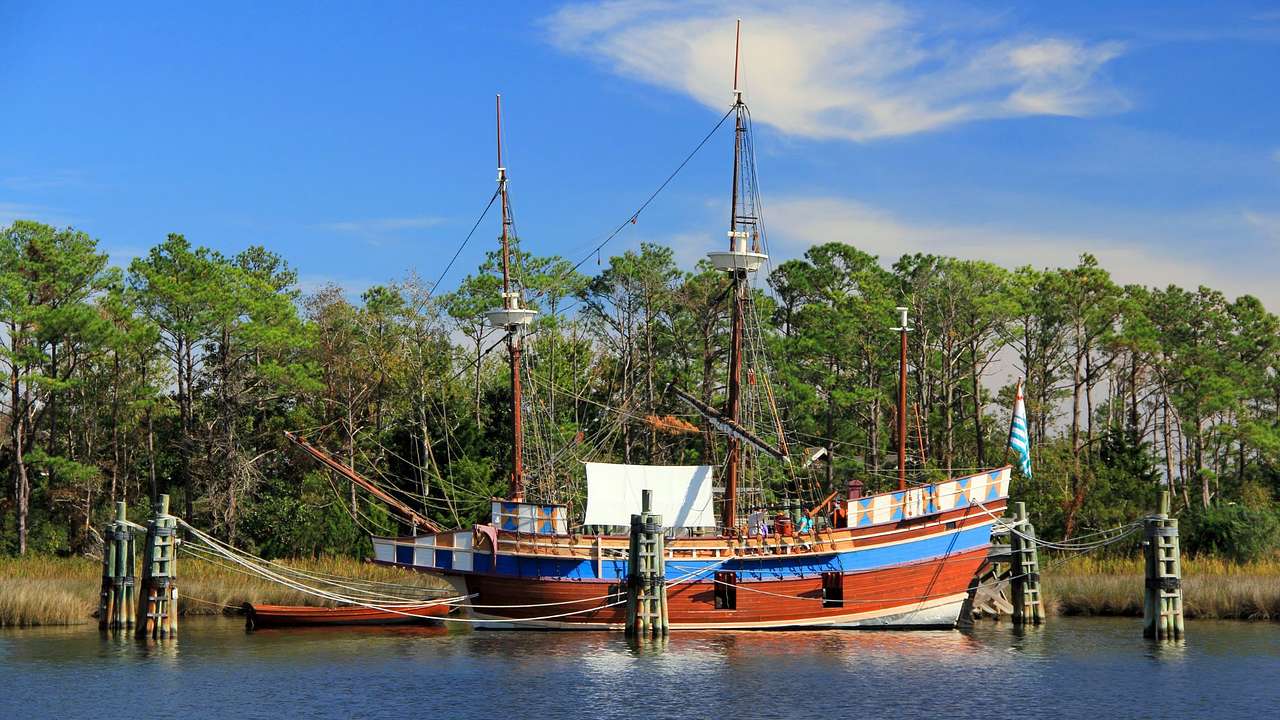 An old-fashioned ship replica on the water with trees behind it under a blue sky