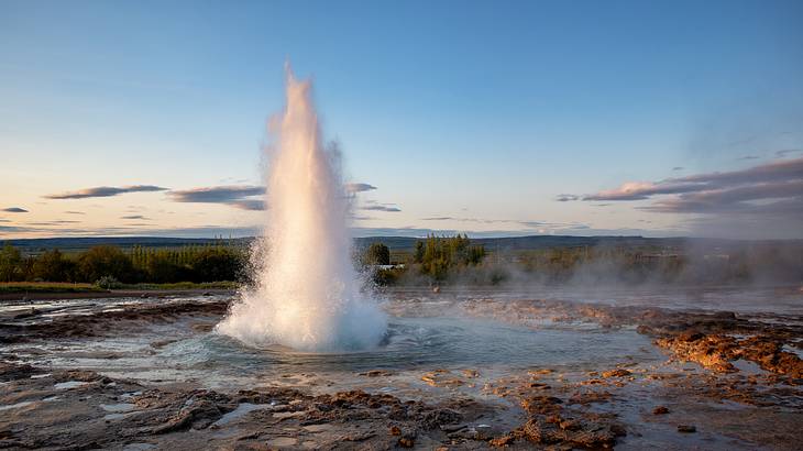 An eruption of water and steam amidst rocky terrain, with greenery in the distance