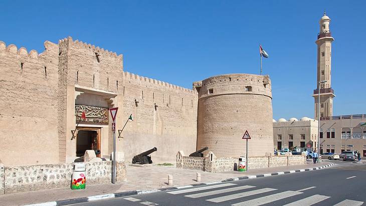 A sandstone fort structure and a tower next to a road on a clear day