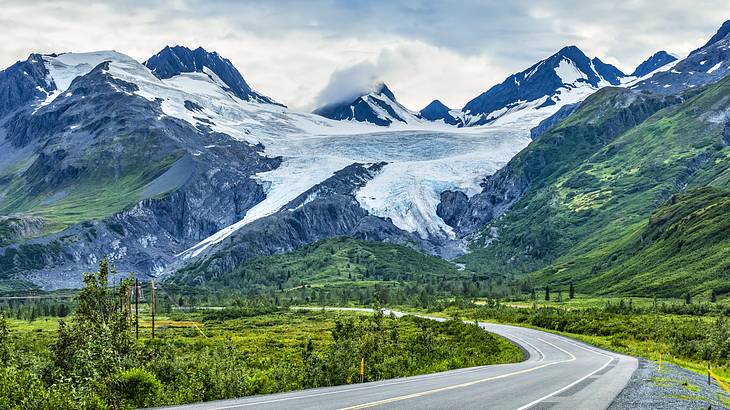 A winding road surrounded by greenery at the foot of a mountain range with a glacier