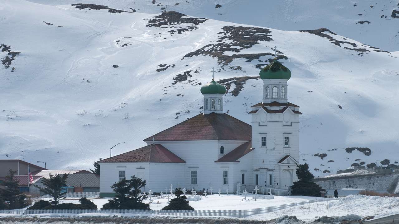 A white church with red roofs and green domes with a snowy mountain in the back