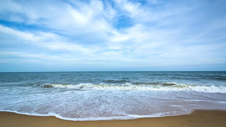 A beach with waves crashing onto the shore under a blue sky with clouds