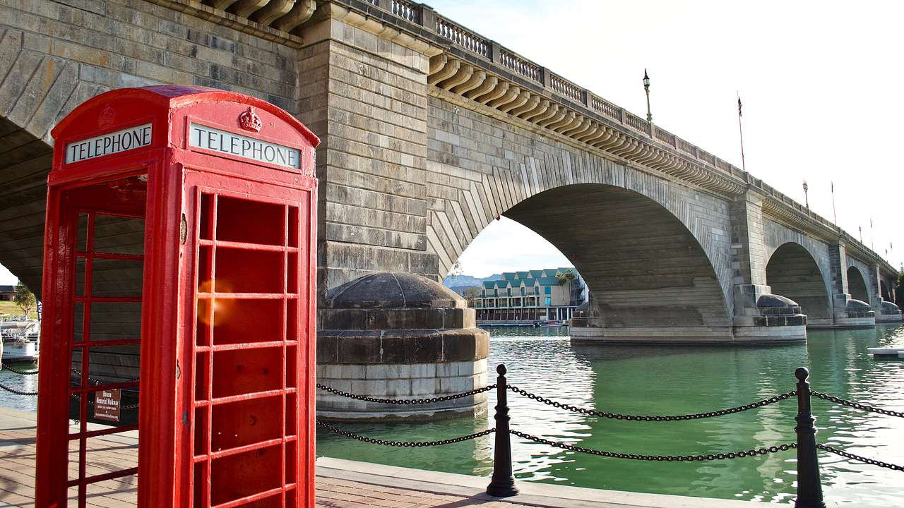 A red English phone booth beside a lake with a granite bridge behind it