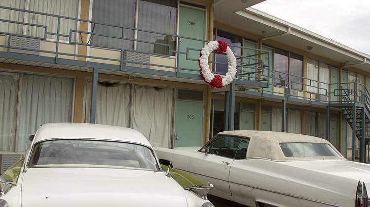 A two-story, older motel with a balcony and two cars parked in front