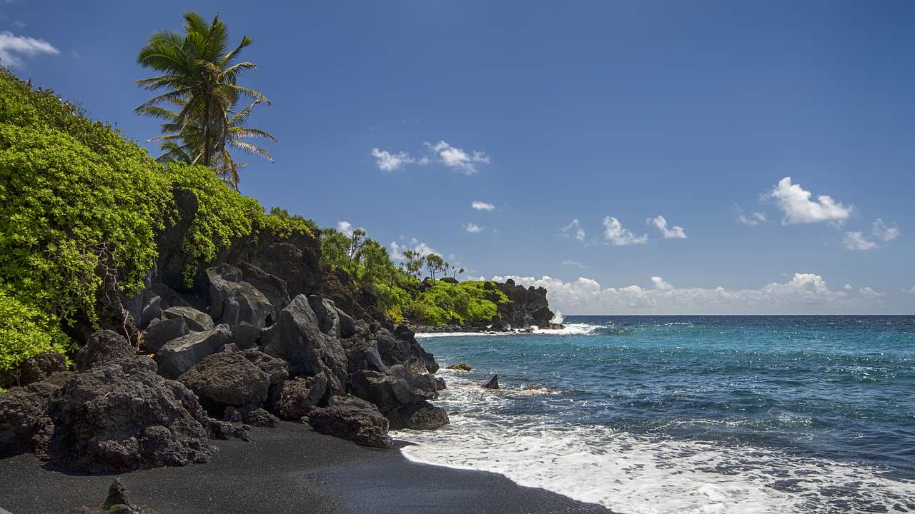 Waves crashing against the black sand of a beach with large rocks and greenery