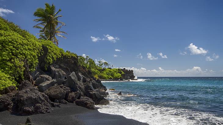 Waves crashing against the black sand of a beach with large rocks and greenery