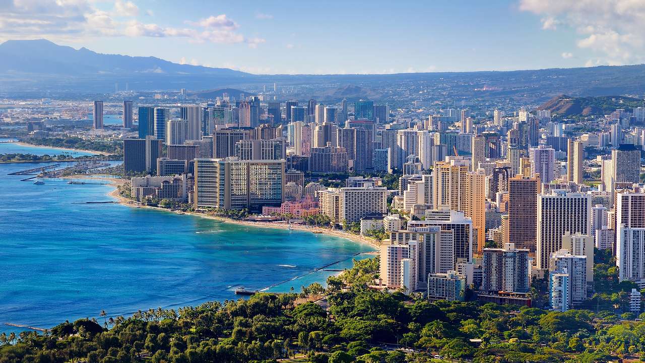 Aerial shot of a coastal city with buildings, greenery, and a mountain at the back