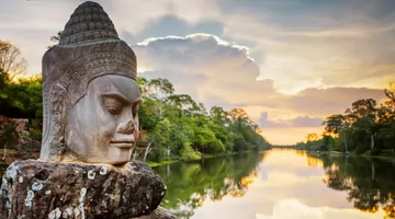 Une statue à face de pierre sur un sentier surplombant l'eau et les arbres au coucher du soleil 