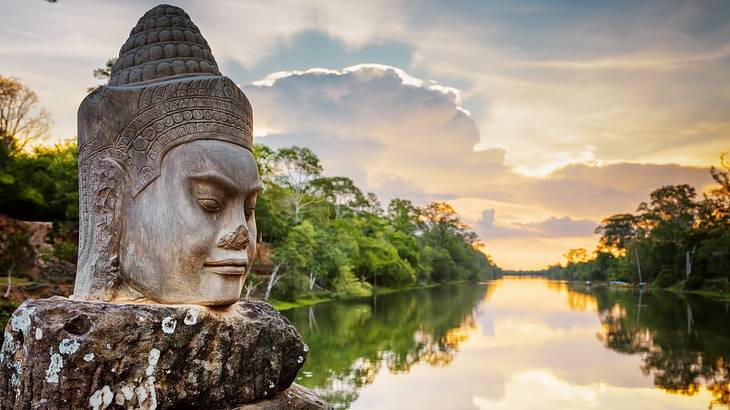 A stone-faced statue on a pathway overlooking water and trees at sunset A stone-faced statue on a pathway overlooking water and trees at sunset