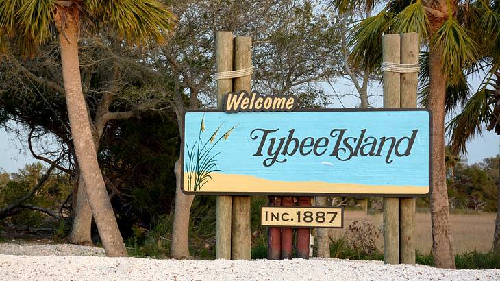 A sign that says Tybee Island on the sand with palm trees next to it