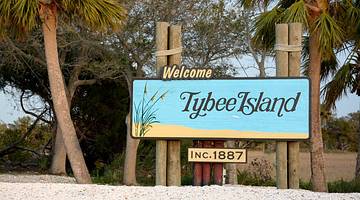 A sign that says Tybee Island on the sand with palm trees next to it