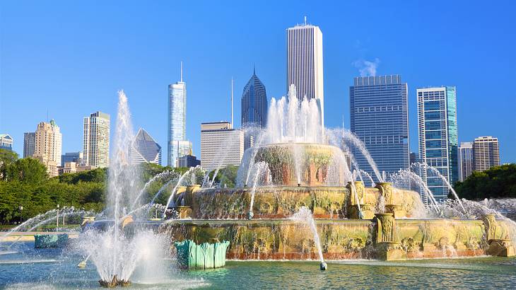 A large water fountain with skyscrapers behind it under a blue sky