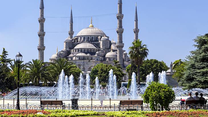 Green plants, flowers, and a water fountain against a mosque with blue domes