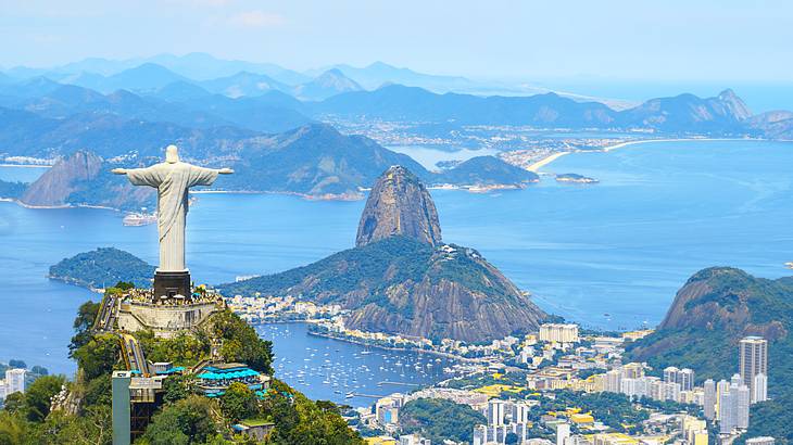 A statue of Jesus on a mountain overlooking buildings, a harbor, and other mountains