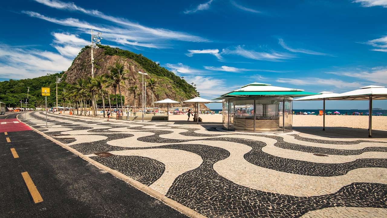 A mosaic sidewalk by a beach with patio umbrellas and palm trees against a green hill