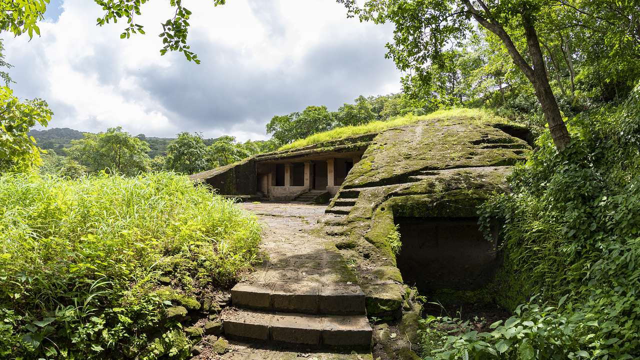 A cave with columns surrounded by greenery and trees under a cloudy sky