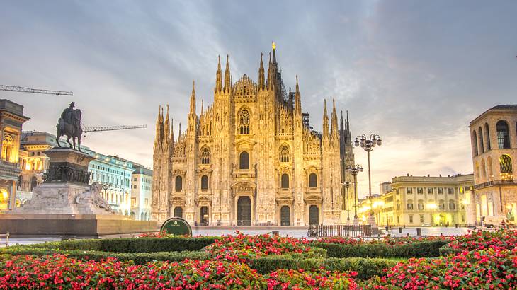 A gothic cathedral building overlooking a statue and a small garden with red flowers