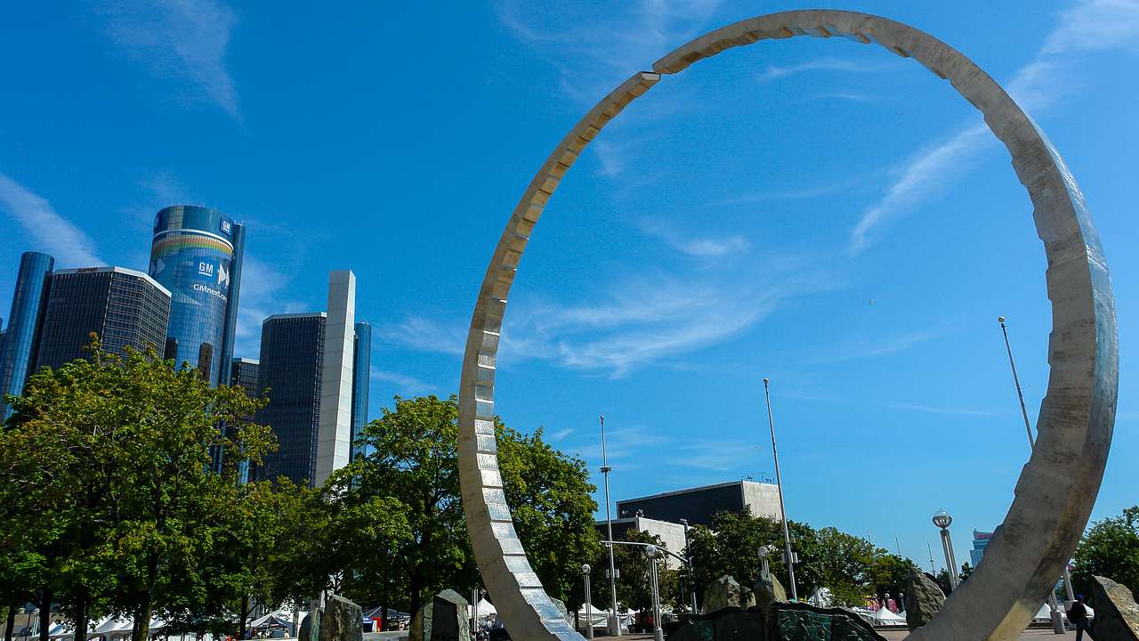 A circular metallic monument surrounded by trees with buildings in the background