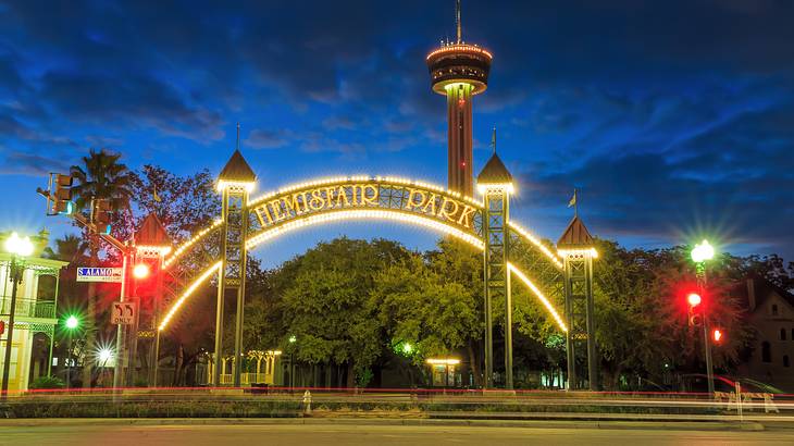 Wide round illuminated gate saying "Hemisfair Park" in front of a sky tower at night