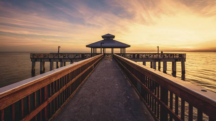 A boardwalk over a sea leading towards a hut-style wooden pier against a sunset sky