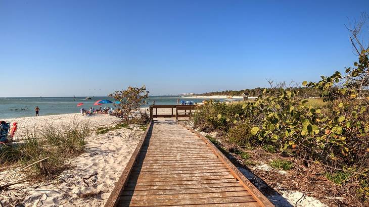 A boardwalk on a white sand beach with some bushes on the right, in front of the sea