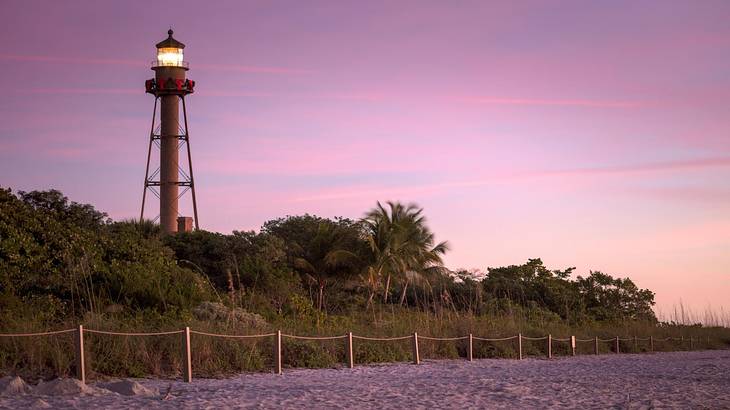 A white sand beach with a lighthouse surrounded by dense trees against a purple sky
